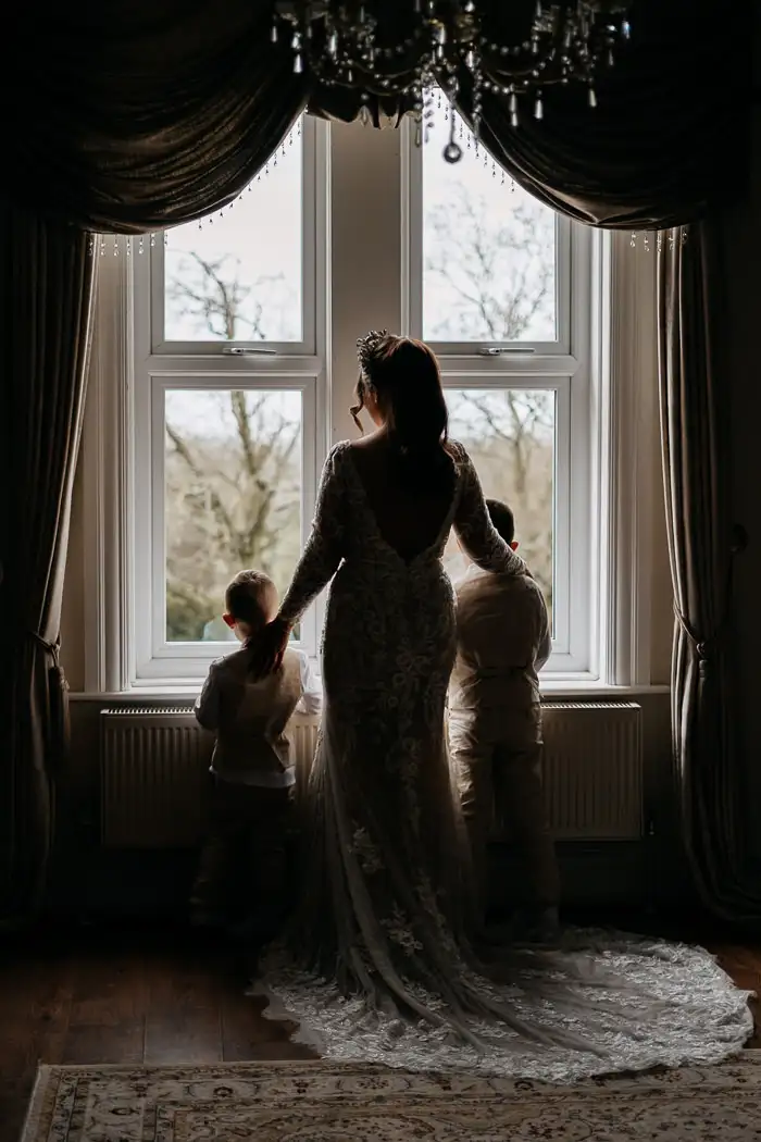 bride stands near a window at west tower wedding venue, with arms around her two page boys